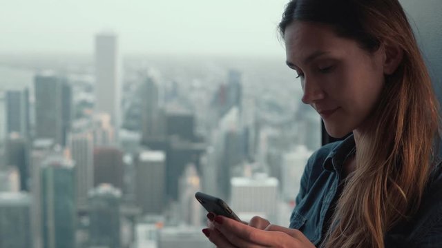Portrait of young attractive woman standing near the window in skyscraper in Chicago, USA and using smartphone.