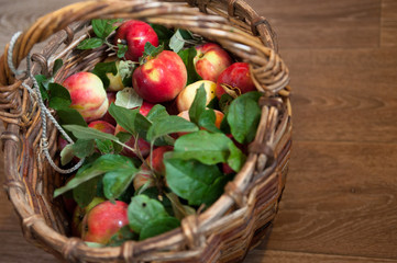 A wicker basket full of red ripe apples with green leaves stands on a wooden table .  the view from...