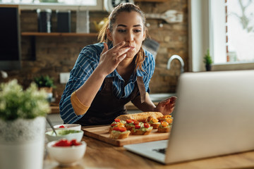Young happy woman using laptop while preparing bruschetta in the kitchen.