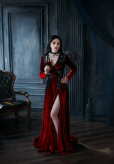 Medieval young woman vampire holds glass of bloody wine in hand. Red long sexy dress train. Gothic...