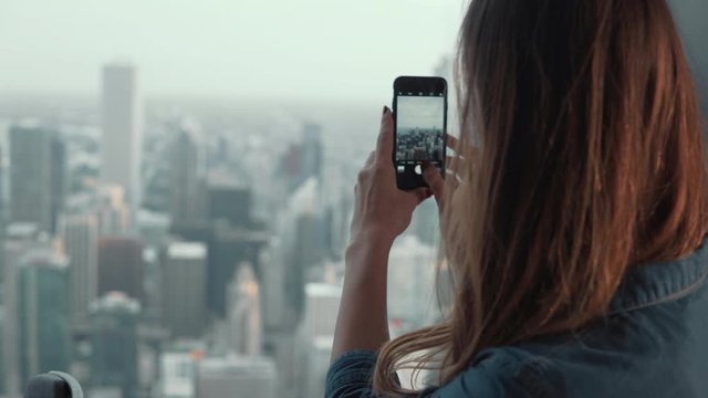 Young attractive woman taking photos on smartphone. Female shoots panorama of Chicago, America from skyscraper.