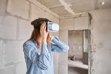 Woman architect or construction engineer wear virtual reality glasses inside a building site. Concrete walls in new apartment, concept of repair.