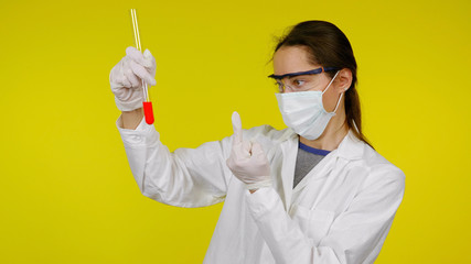 Doctor in a medical mask, goggles and latex gloves shows fuck analysis to the flask. Young girl in a white coat on a yellow background