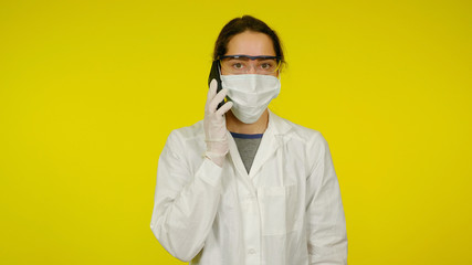 Young doctor in a medical mask, goggles and latex gloves is talking to the patient on the phone. Girl in a white coat on a yellow background holds a smartphone near her ear. Coronavirus, flu