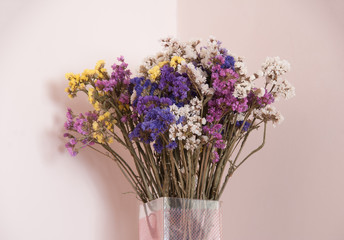 A close up photo of dried up violet, white and yellow flowers in a glass vase. Pink background