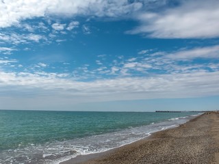 Blue sky with clouds over the green water of the sea and the coast of sand and pebbles.