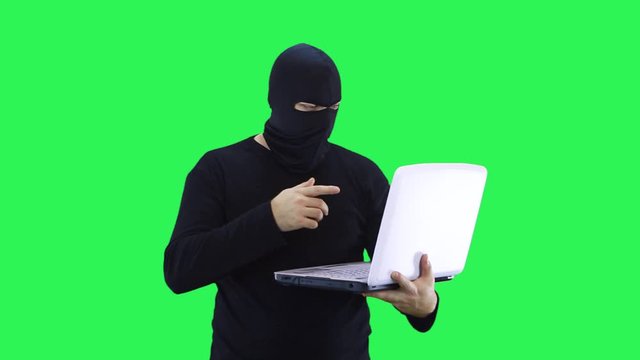 A man in a black mask talks to the interlocutor and enters data into a laptop, on a green background