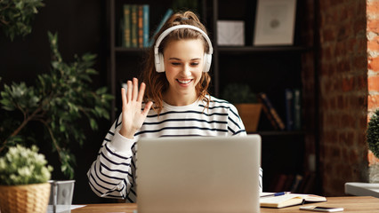 Cheerful woman in headphones greeting friend while talking on laptop at home. © JenkoAtaman