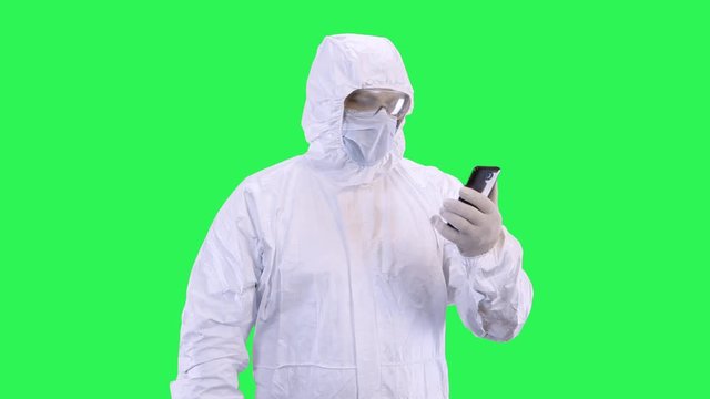 A man in a mask and a protective suit is talking via video calling on a smartphone while standing against a green background.