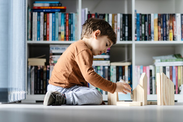 full length view of small caucasian boy little child ar home kid playing with wooden toys brick in shape of house sitting on the wooden or vinyl laminated floor alone developing creativity side view