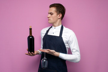 young guy sommelier holds a bottle of red wine on a colored background, an expert on wine in a...