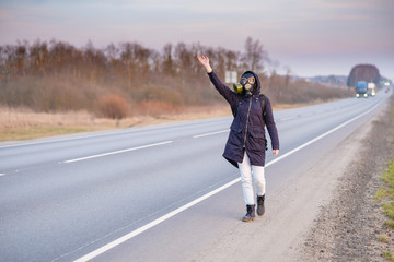 Obraz premium COVID-19. A girl in dark clothes, a gas mask and a hood on her head. Cloudy day in the country. She raises her hand and tries to stop the passing car. Escaping from a zone infected with a coronavirus.