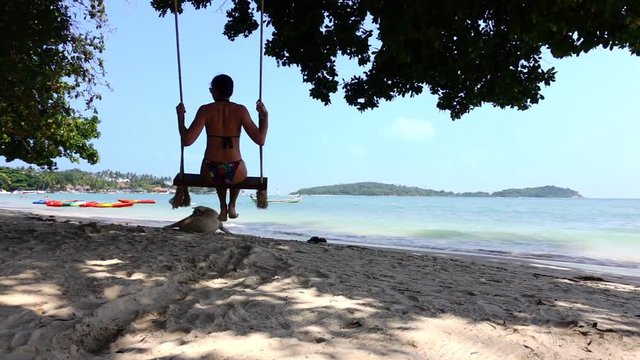 Young woman is swinging on swings at the seashore. Slow motion of teeting woman. Theme of vacation, travelling and journey. Sea water is bright and clear and the sun shines at the daytime.