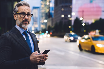 Adult businessman holding smartphone and looking away against New York road