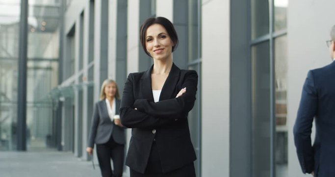 Portrait shot of Caucasian youg beautiful businesswoman standing outdoor and looking straight to camera. Female smiling at street near modern building. Attractive woman in business style.