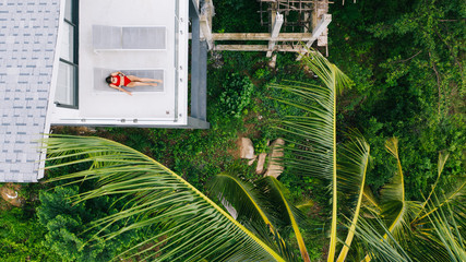 Aerial top view, female tourist in red swimsuit lying on sun bed in contemporary villa surrounded by tropical palm trees. Property rental during summer vacations holidays. Enjoying rest and relaxation