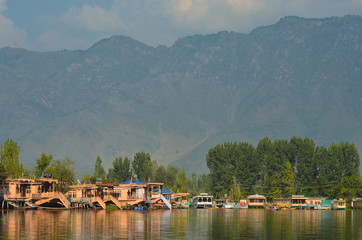 Fototapeta na wymiar Wooden houseboats moored on Dal Lake in Kashmir. Their colours are reflected in the water. Trees line the shore, and mountains rise behind them to a cloudy sky.