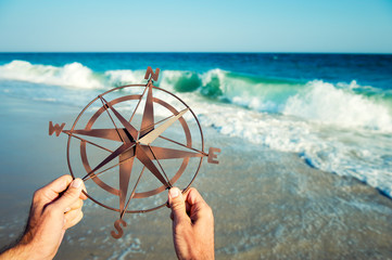 Hands of unrecognizable man holding simple metal compass rose outdoors in front of shoreline with crashing waves