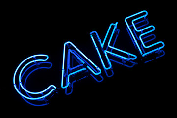 Nighttime view of all-caps CAKE message glowing in blue diagonal letters in neon on black background