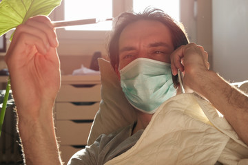 Coronavirus quarantine: young man lying in his bed with medical mask and thermometer
