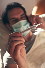 Coronavirus quarantine: young man lying in his bed with medical mask and thermometer