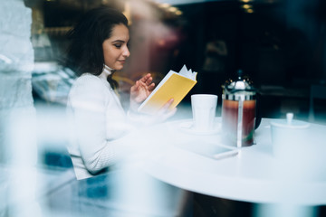 Carefree woman enjoying time for reading romantic novel during leisure day in coffee shop, attractive hipster reader sitting at cafeteria table with hot brewed tea and recreating with literature