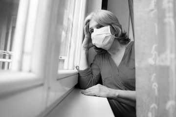 Sad senior woman wearing surgical mask looking through the window, covid19 pandemic 