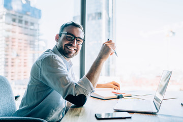 Portrait of cheerful male entrepreneur in classic eyewear smiling at camera while working at...