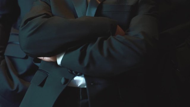 Groom dressed in black tuxedo with bow tie and white floral boutonniere is crossing hands on the chest, front view, wedding day, formal attire