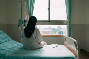 back view of lonely young asian woman sitting on bed in hospital ward. depressed female patient stay in room with body check result paper beside. sad lady in pajamas looking out window indoor