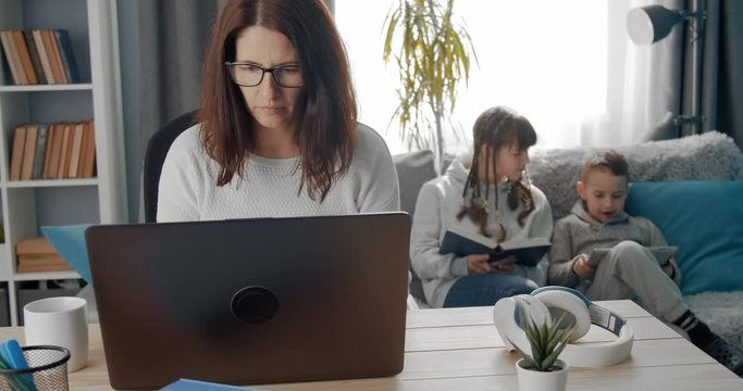 Concentrated female freelancer in eyeglasses working on laptop at home and parenting her two children during quarantine. Remote female business, career. Lifestyle moment.