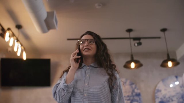 Low angled shot of young beautiful woman in eyeglasses walking through cafe and speaking on mobile phone