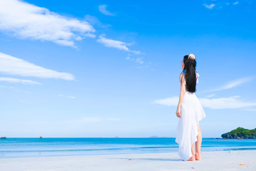 Fototapeta na wymiar Asian woman, long black hair, wore white dress and hat standing on the beach near the woven bags and sunglasses and facing back by the sea with copy space blue sky,summer holiday and vacation concept