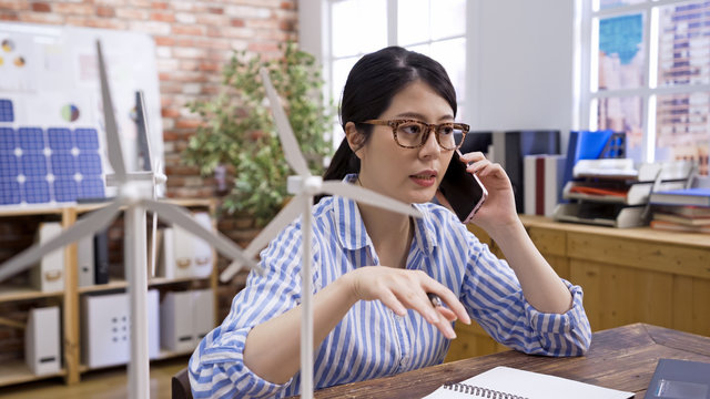 small business startup and people concept. elegant female engineer worker calling on smartphone in office. young girl architect employee discussing wind energy building on cellphone in workplace.