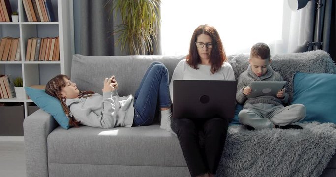 Portrait of happy family sitting on grey couch with personal gadgets. Mother and her children spending time at home with modern technology.