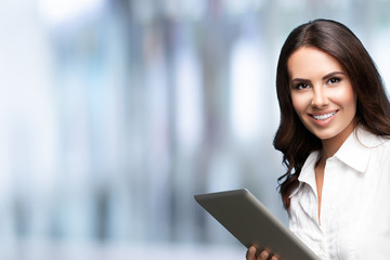 Happy smiling beautiful young brunette businesswoman using no-name tablet pc, against blurred office background, with copy space for some slogan or text. Success in business concept.