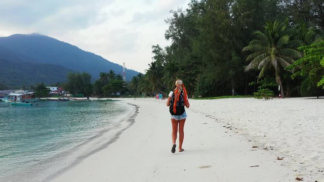 Stylish young girl on short and flip flops walking slowly on white sand of exotic beach washed by calm turquoise lagoon in Myanmar