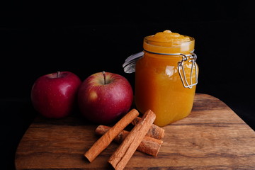 Home made apple jam in a mason jar. Red apples and cinnamon on a wooden platform. Black background. 
