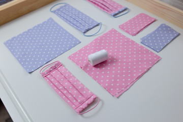 Face mask against virus handmade, sew protective mask, pieces of dotted cloth, threads on white table. Flat lay with pink and blue cloth, scissors and threads.