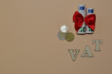 Word VAT made of wooden letters and  50 and 10 euro banknotes, few coins, in rolls tied with red ribbon bow top view on pastel color background copy space