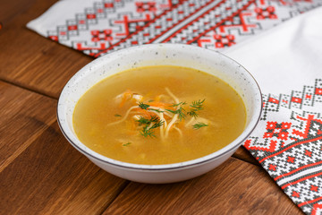 Chicken soup with noodles and vegetables served in white bowl over rustic wooden table with traditional tablecloth.