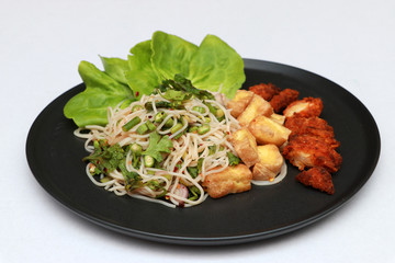 Breakfast in the black round plate on white floor. Rice vermicelli spicy salad with Fried Tofu, Fried chicken and lettuce vegetable.