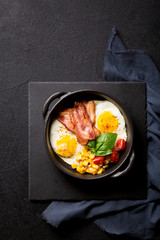 Fried egg with slices of bacon, tomato, corn and spinach black frying pan on a black background. Breakfast. Top view