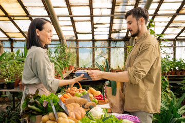 Young bearded man in casual shirt paying with contactless card for fresh organic products at farmers market