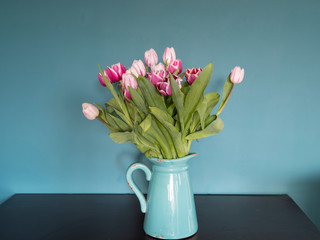Fototapeta premium Fresh pink and red tulip flowers bouquet in a jug on a brown table in front of green wall. View with copy space
