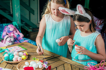 Cute little girls are painting Easter eggs with brushes staying next to the wooden table in backyard.