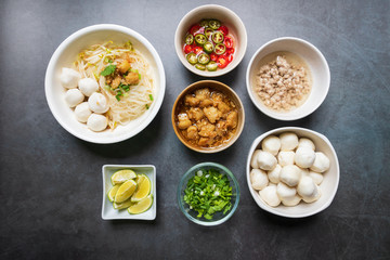 Rice stick noodles with fish ball in white bowl. Garlic cracklings with lard, mined pork, pickled chili, lime and coriander with spring onion various ingredients and condiment on table.