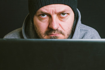 Man wearing black cap and hoodie sitting behind the computer monitor in the dark, looking to camera, close up, hacking computer system concept
