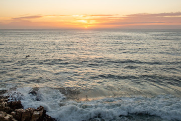 Green hued waves crashing on rocks with beautiful golden clouds at sunrise on the horizon.