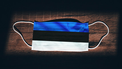 Estonia National Flag at medical, surgical, protection mask on black wooden background. Coronavirus Covid–19, Prevent infection, illness or flu. State of Emergency, Lockdown